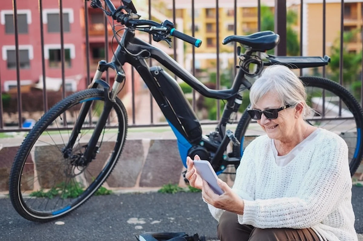 Starsza kobieta siedząca na zewnątrz obok roweru elektrycznego, używająca telefonu w rozmowie wideo